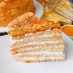 The Best Russian Honey Cake sliced cake on a white platter with a cake in background with a yellow and white dotted towl , whisk and honey in a white container on a black tabletop.