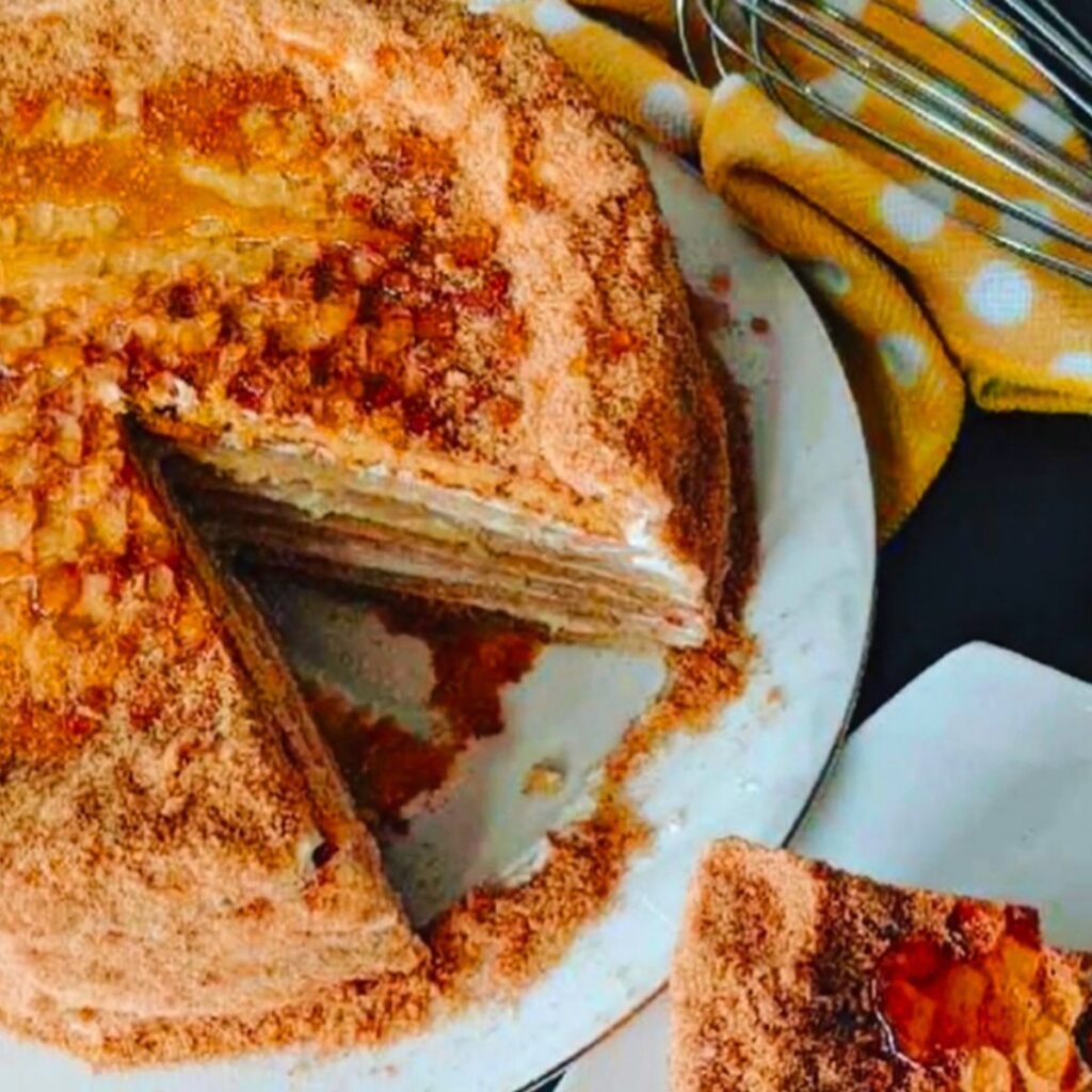 The Best Russian Honey Cake on a white cake platter with a yellow white dotted towel, whisk and a cut sliced cake with honey in a small saucer on a black tabletop.