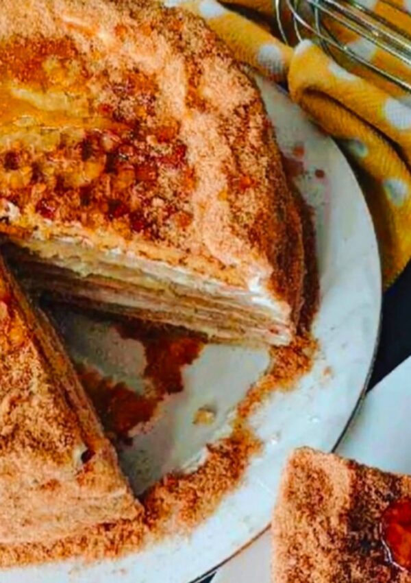 The Best Russian Honey Cake on a white cake platter with a yellow white dotted towel, whisk and a cut sliced cake with honey in a small saucer on a black tabletop.