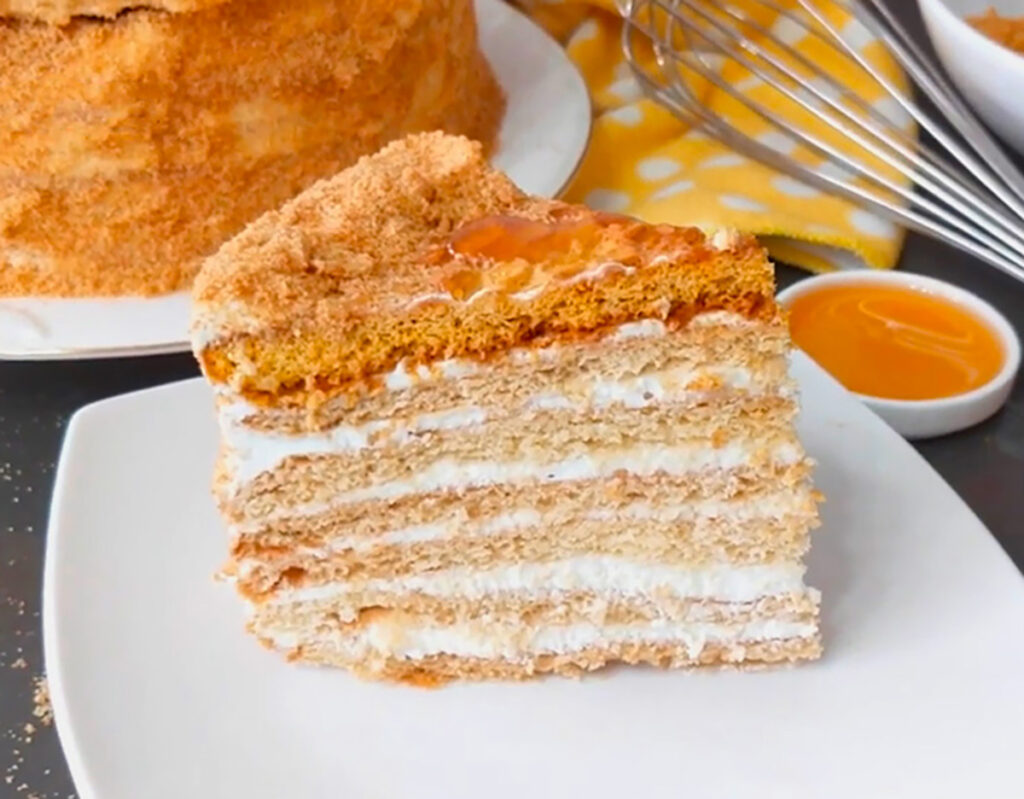 The Best Rusian Honey Cake sliced cake on a white platter with a cake in background with a yellow and white dotted towl , whisk and honey in a white container on a black tabletop.