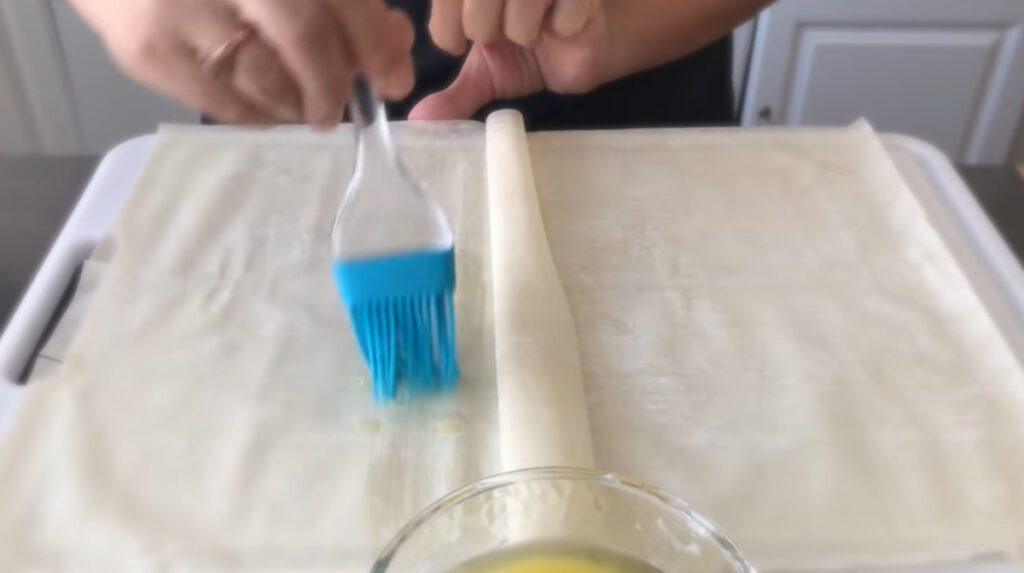 Spinach Feta Borek - sheets of phyllo dough on top of the table brushed with blue rubber baking brush melted butter over each ply of dough with a glass bowl of melted butter inside. Two hands in the picture with a person in the background with a black shirt.