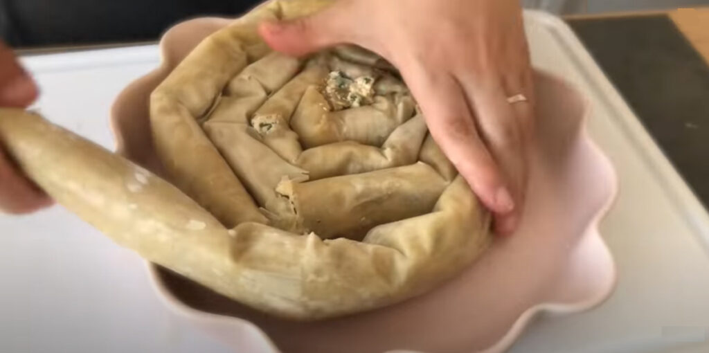 Spinach Feta Borek rolling filled dough into a spiral with two hands with someone in the background with a black shirt in a pink pie baking pan.