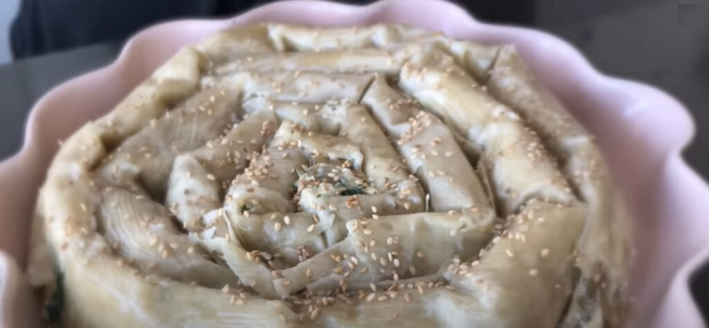 Spinach Feta borek spiral unbaked with roasted sesame seeds sprinkled on top of phyllo dough in a pink pie baking pan.