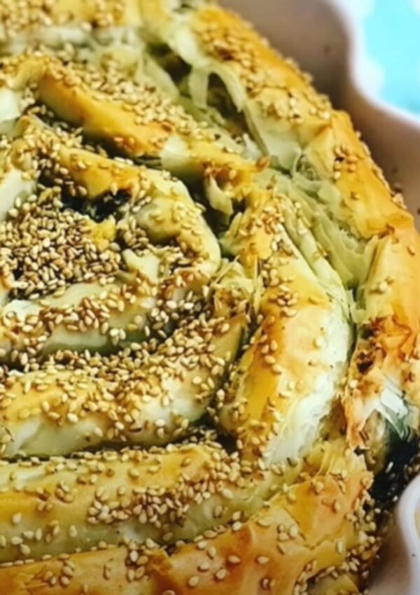 Spinach Feta Borek spiral baked with roasted sesame seeds sprinkled on top of baked phyllo dough in a pink pie baking pan with a teal blue towel and wooden chopping board on the tabletop.