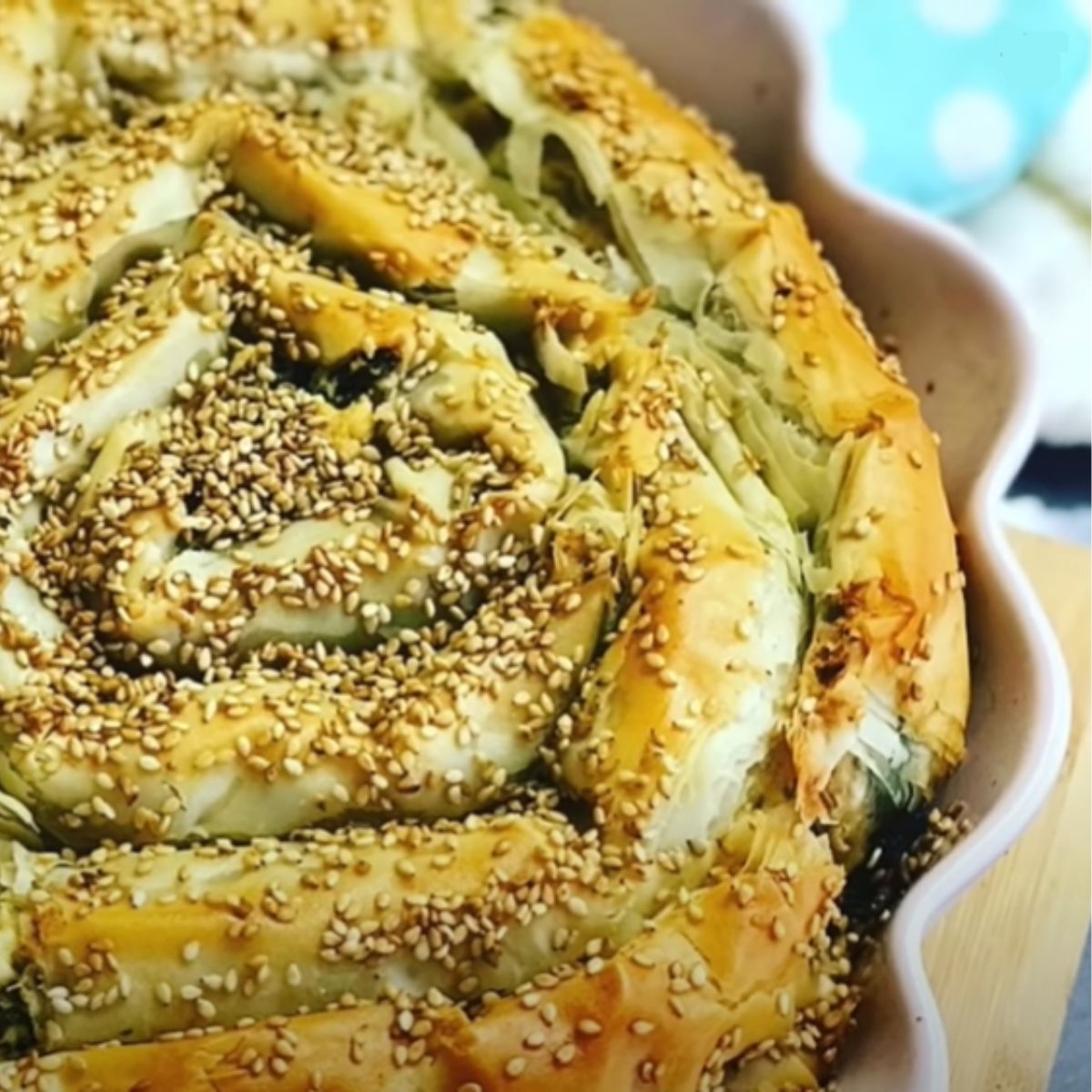 Spinach Feta Borek spiral baked with roasted sesame seeds sprinkled on top of baked phyllo dough in a pink pie baking pan with a teal blue towel and wooden chopping board on the tabletop.
