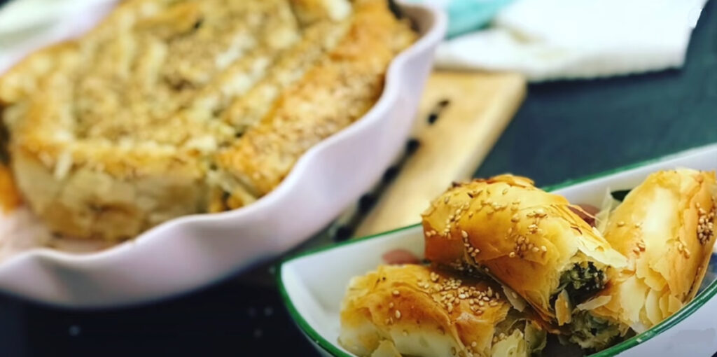 Spinach Feta Borek spiral baked with roasted sesame seeds sprinkled on top of baked phyllo dough in a pink pie baking pan with a wooden coaster underneath baking pan, teal mitten in background with cut pieces of spinach feta borek placed in a white dish with a green trip around the dish.