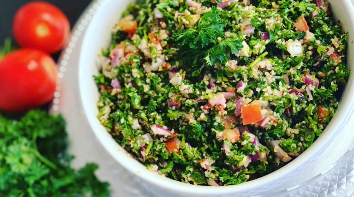 Tabbouleh - Healthy Salad in a white bowl accented with a silver platter under a white bowl accented with parsley on a black countertop