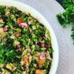 Tabbouleh - Healthy Salad in a white bowl accented with a silver platter under a white bowl accented with parsley on a black countertop with tomatoes and parley to the right.