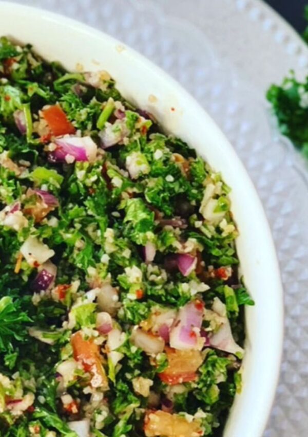 Tabbouleh - Healthy Salad in a white bowl accented with a silver platter under a white bowl accented with parsley on a black countertop with tomatoes and parley to the right.