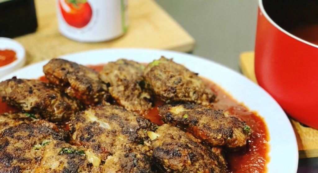 Beef Cutlets in Tomato Marinara Sauce on a white plate with tomato sauce at the bottom with a wooden chopping board and italian seasoning sprinkled on the background.
