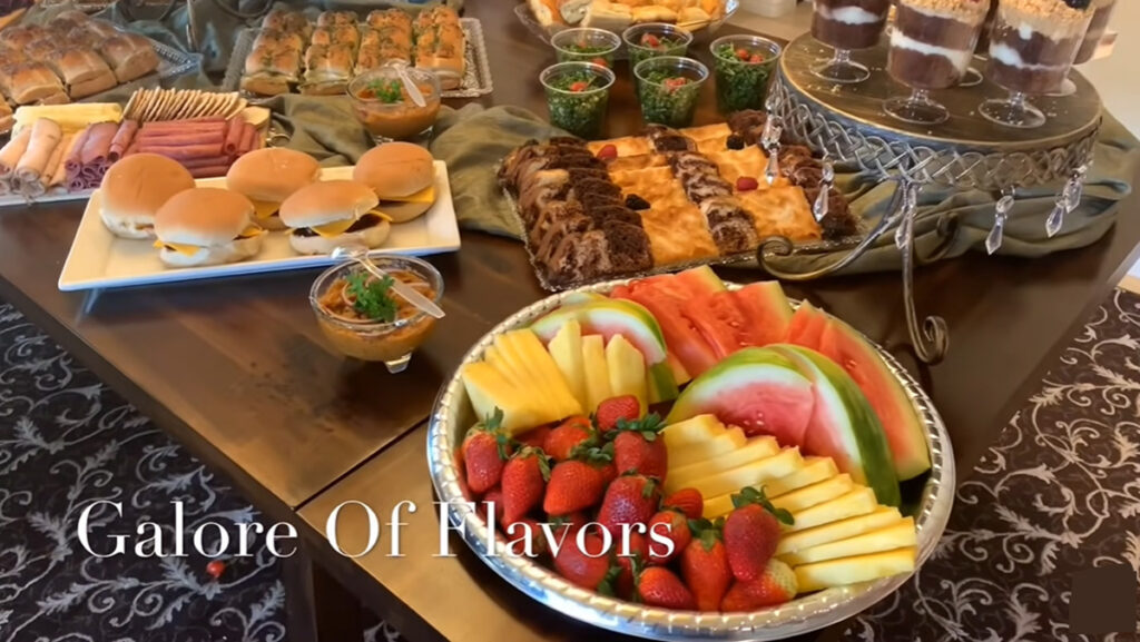 The Best Party Buffet Food Recipe table setup fruit displayed on a round silver platter, a white plate with cheese burgers, assorted cakes in a crystal dish, salad cups, and pesto basil buns in a crystal dish with green accent fabric on a dark wooden table.