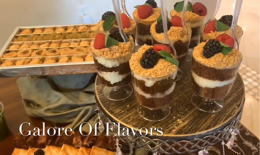 The Best Party Buffet Food Recipe pudding chochlate cake cups with a cookie crumble topping and fresh fruit accented to mint leaves, assorted baklava platter in the background, assorted cakes on platter below pudding cups.