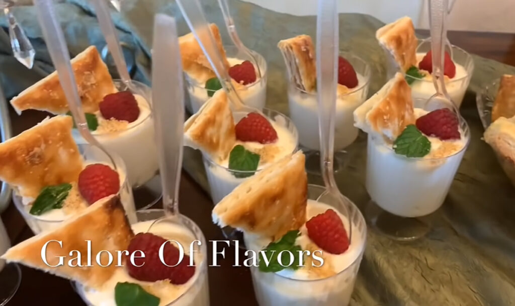 The Best Party Buffet Food Recipe pudding cups with fresh rasberries ontop with a puff pastry sweet waffer accented with a fresh mint leaf with plastic forks submerged inside the cup with a green accent fabric draping wooden table.