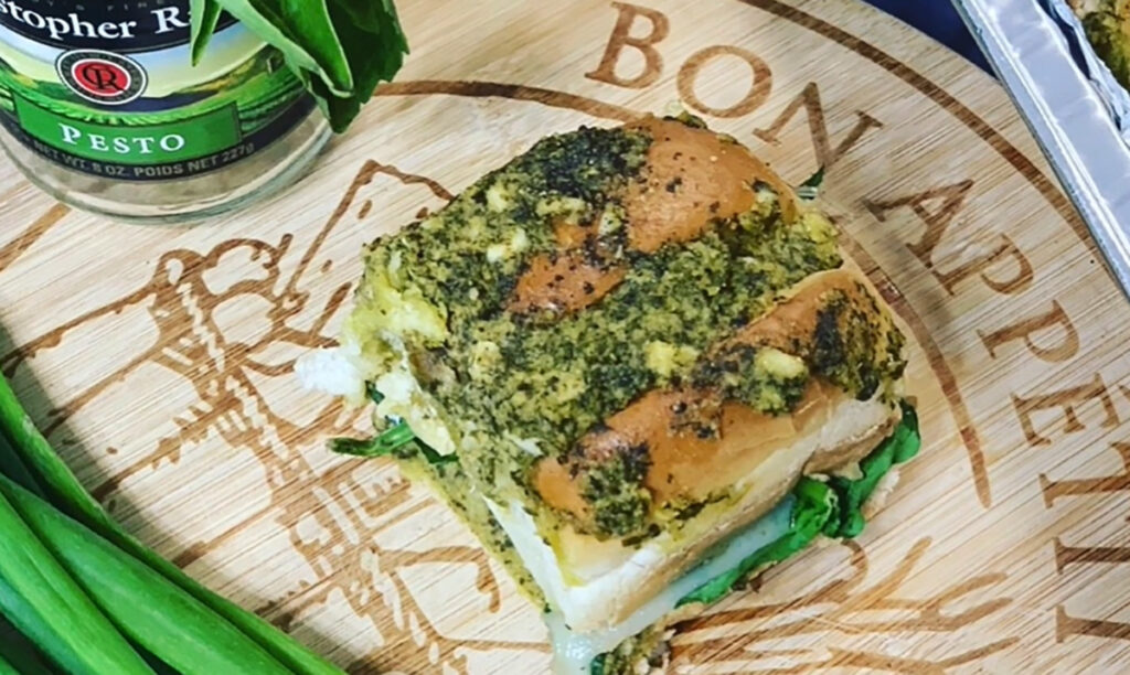 Basil Pesto Swiss bite on a wooden chopping board that says Bon Appitite with a carved wooden board with fresh basil in a jar in the background.