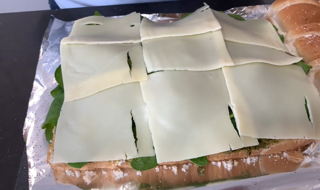 Hawaiian rolls are cut in half on a piece of foil and pesto and spinach leaves and sliced swiss cheese added ontop of a black tabletop.