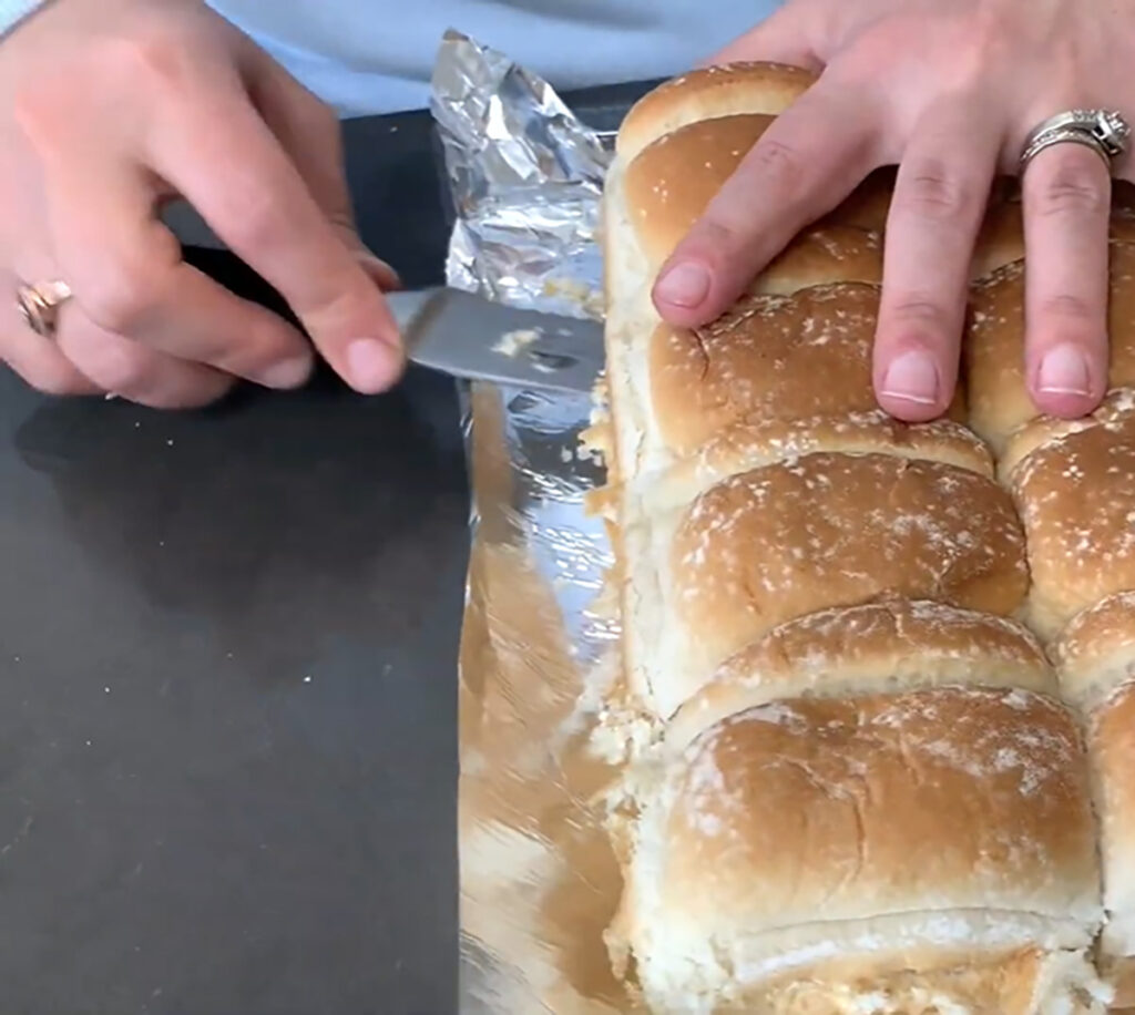 Hawaiian rolls on a piece of foil cutting through with a bread knife on a black tabletop.