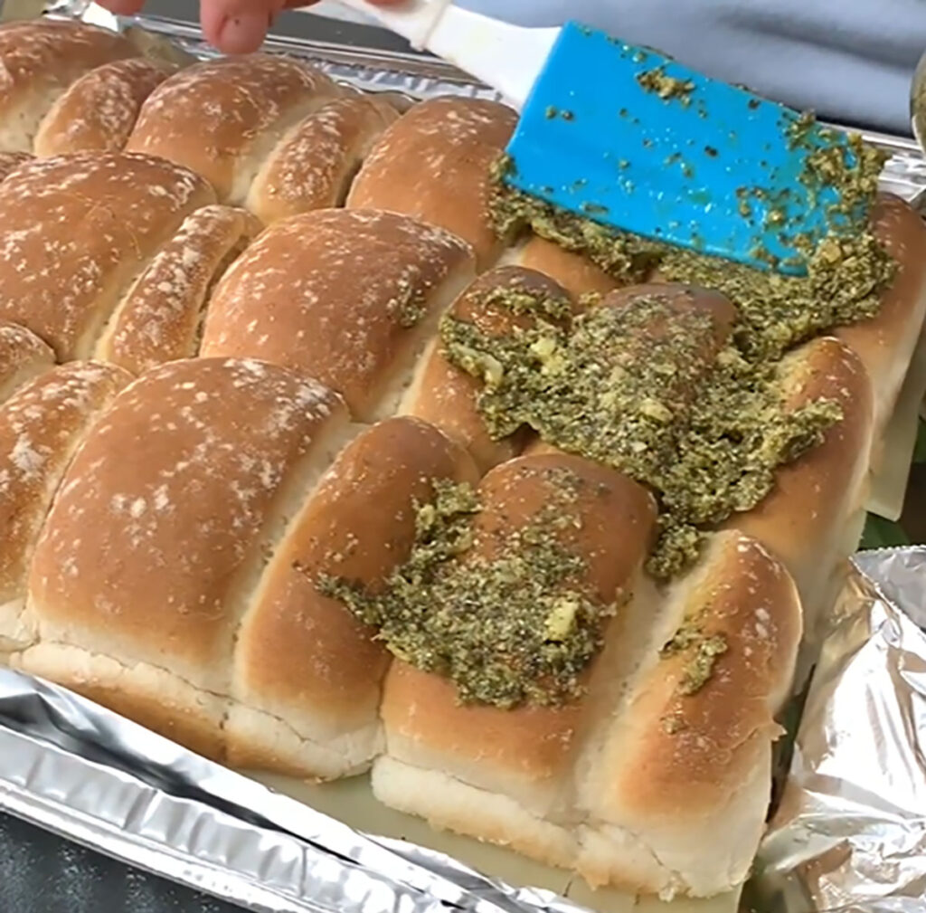 Hawaiian rolls spreading pesto sauce over top of bread buns in a foil tin baking pan with a blue rubber spatula on a black tabletop