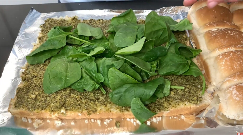 Hawaiian rolls are cut in half on a piece of foil and pesto and spinach leaves added ontop of a black tabletop.