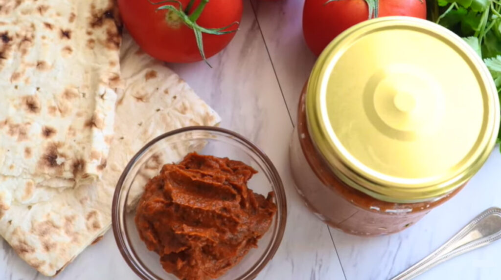 Roasted eggplant spread in a glass small bowl with a sterilized jar filled with spread with a gold lid with lavash and tomatoes laying on top of a white countertop.