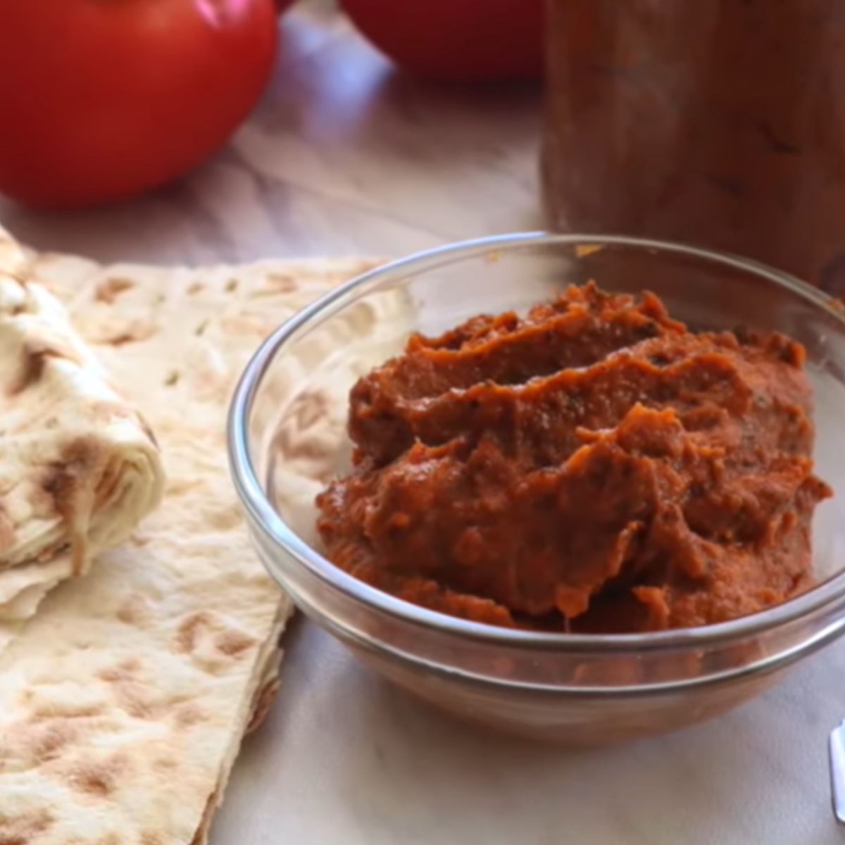 Roasted vegetable spread in a small glass bowl with lavash bread lying on a white marble countertop with tomato and a jar filled with eggplant spread.