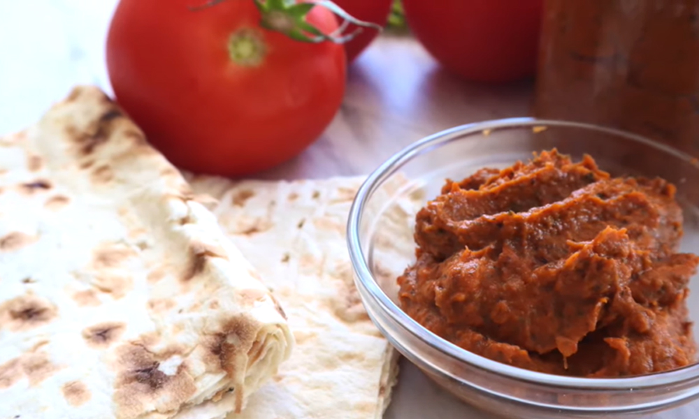 Roasted vegetable spread in a small glass bowl with lavash bread lying on a white marble countertop with tomato and a jar filled with eggplant spread.