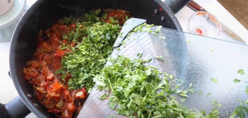 Sauting vegetables in a black saute pan adding chopped cilantro on a glass chopping board.