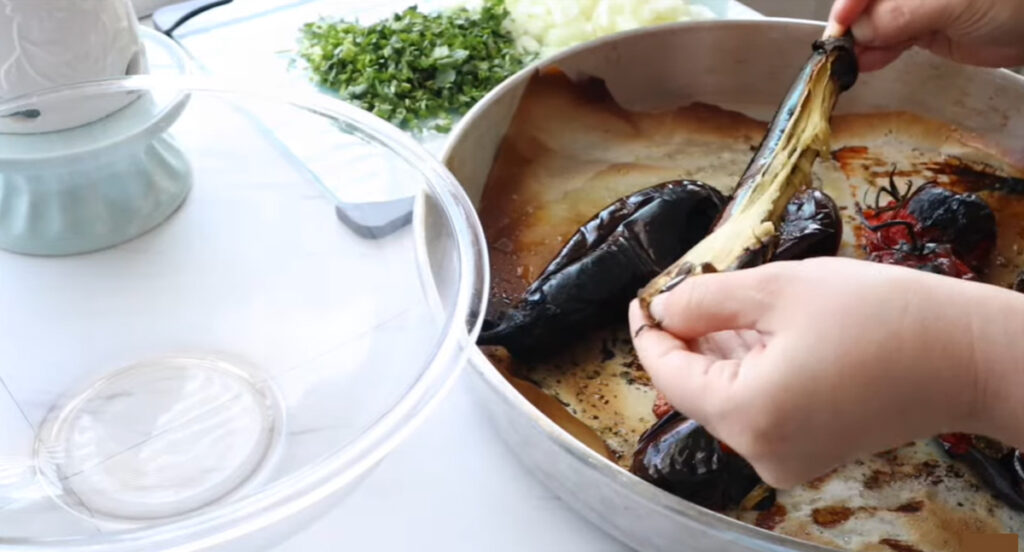 Pealing roasted eggplants in a silver tin baking round pan with a glass mixing bowl and chopping board of chopped cilantro and onions in the background on a white countertop.
