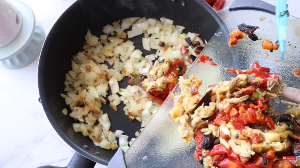 Sauted onions in a black pan placing roasted vegetables in the pan along with onions to saute on a glass chopping board.
