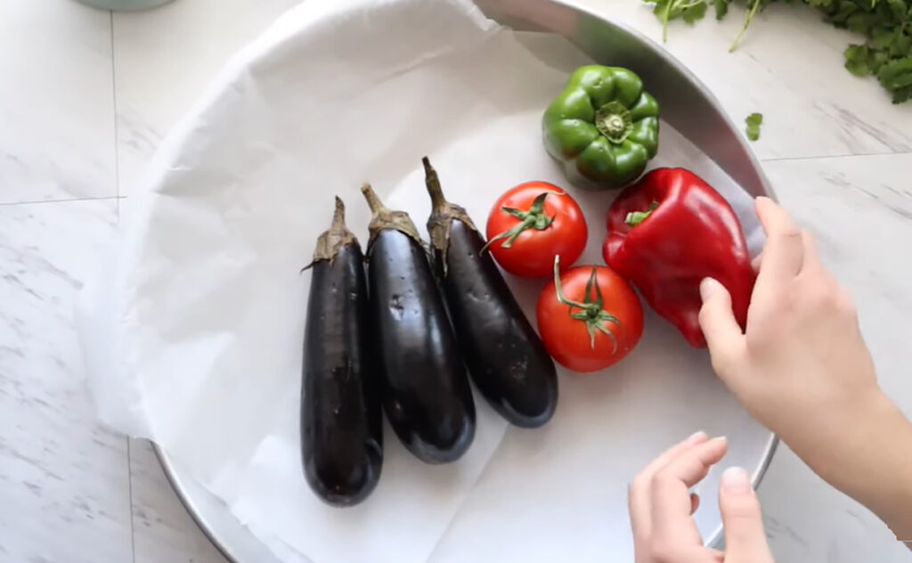 Eggplants, tomatoe, bell peppers in a round baking pan with parchment paper assorting and arranging vegetables on platter with hands.