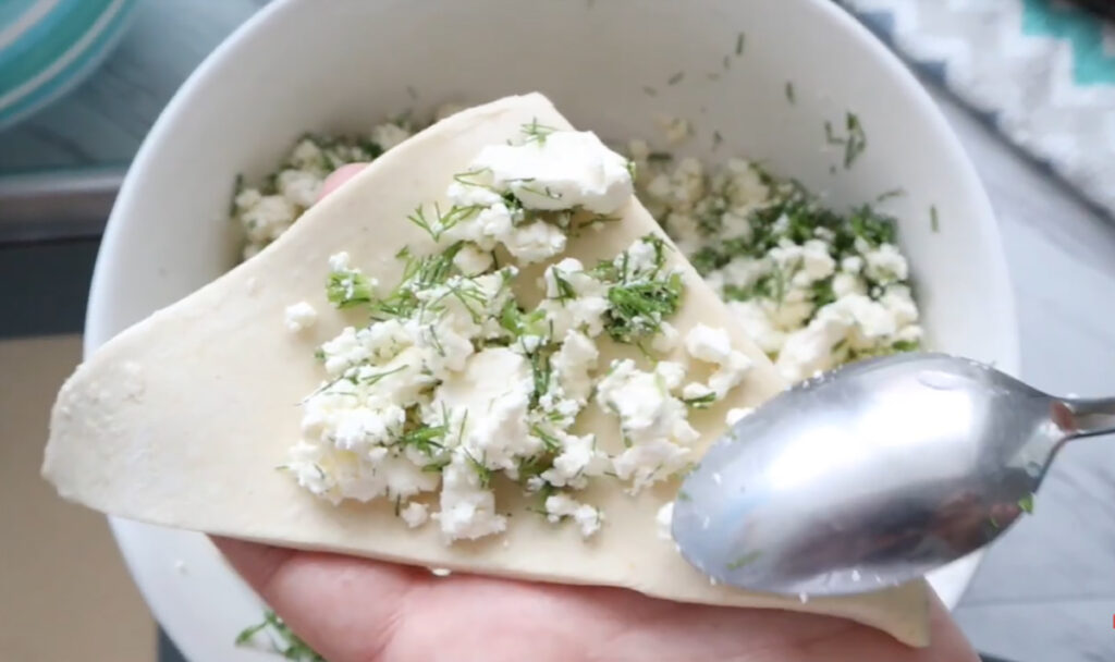 Filling Cheese ontop of puff pastry dough with spoon with a round bowl underneath on the tabletop.