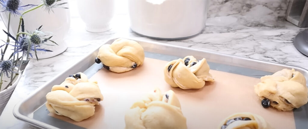 Blueberry Danish Twists before baked lined up on a baking pan on a silicon baking liner with white canisters in the background ontop of a white countertop with blue thistle flour in the background.