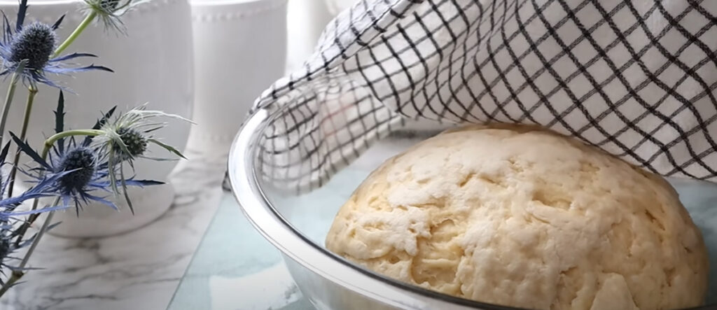 Blueberry Danish Twists glass mixing bowl with dough that has risen with a white and black lined hand towel uncovering dough with white canisters in the background on a white countertop with blue thistle flowers in a vase in the background.