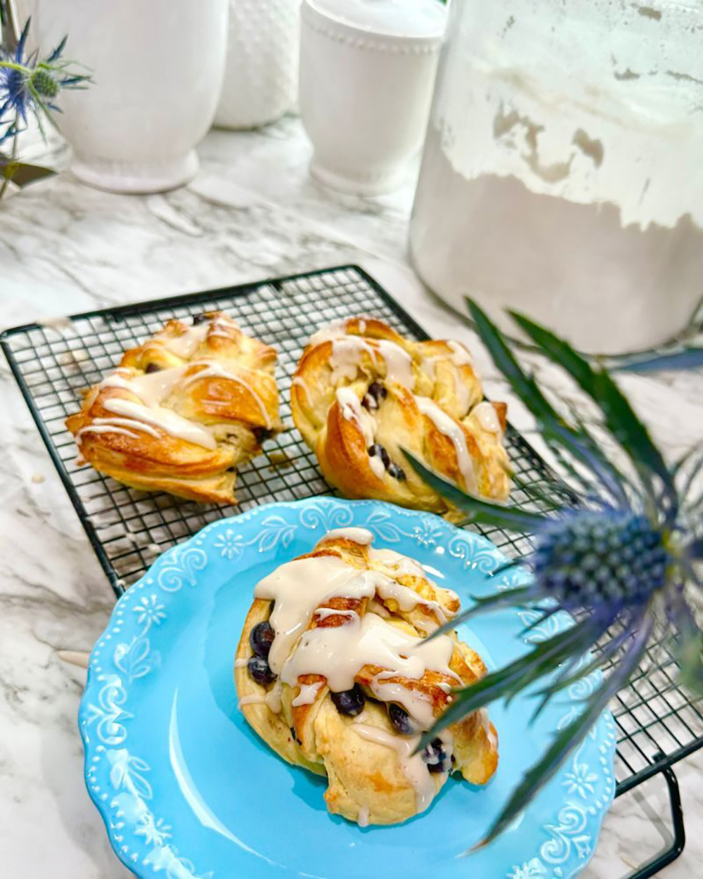 Blueberry Danish Twist Brioche Bread Dessert on a black cooling rack on a white marble counter with white canisters with a canister full of flour accented with blue thistle flower.