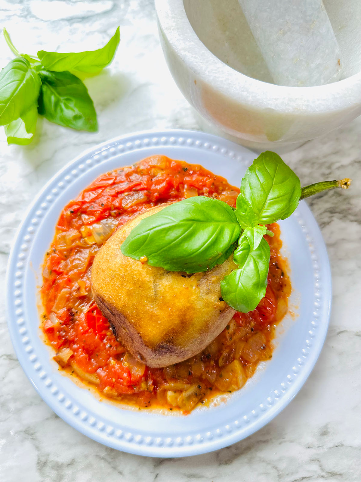 Cheesy Stuffed Eggplant in a Zesty Garlic Tomato Sauce on a white plate accented with fresh basil leaf on top of eggplant with a white stone mortar smasher, fresh basil on a white countertop.