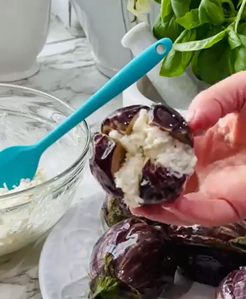 Cheesy Stuffed Eggplant in a Zesty Garlic Tomato Sauce, stuffed eggplants with cheese on a white plate with a white mortar grinder next to a glass bowl with cheese and a blue spatula, fresh basil with two white canisters in the background on a white countertop