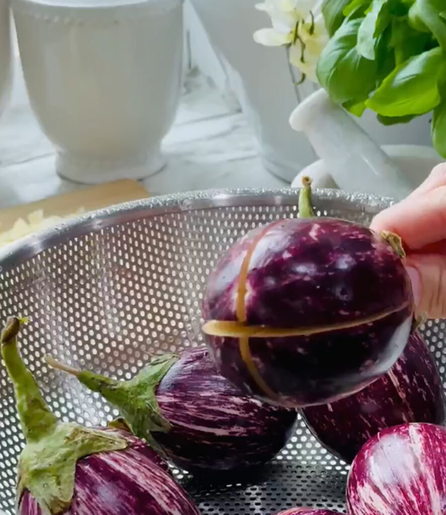 Cheesy Stuffed Eggplant in a Zesty Garlic Tomato Sauce with a white mortar grinder in the background with cut eggplants in a stainless steel strainer with fresh basil with two while canisters on a white countertop