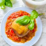 Cheesy Stuffed Eggplant in a Zesty Garlic Tomato Sauce on a white plate accented with fresh basil leaf on top of eggplant with a white stone mortar smasher, fresh basil on a white countertop.