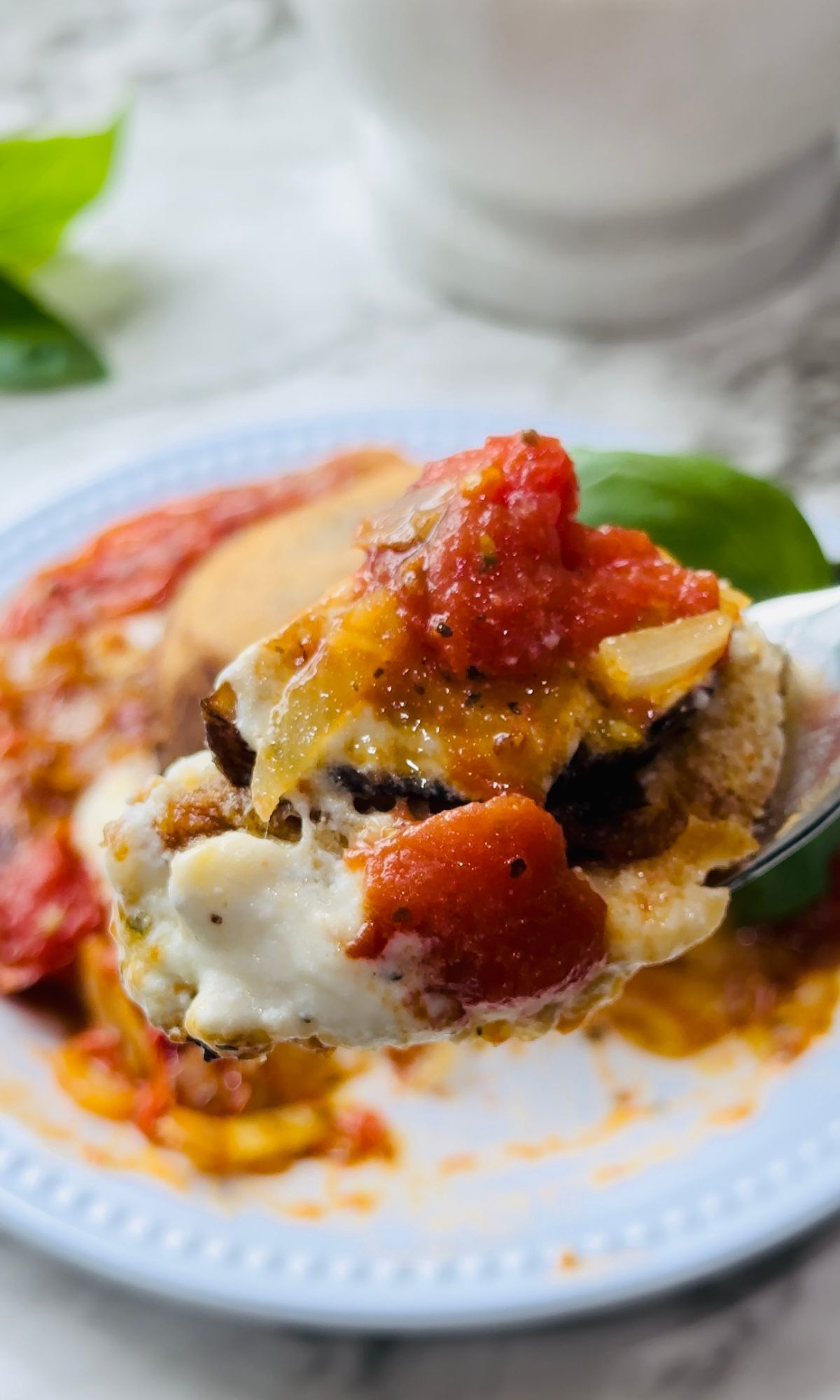 Cheesy Stuffed Eggplant on a white plate with a spoon of a bite-size serving with fresh basil in the background with a white marble countertop.