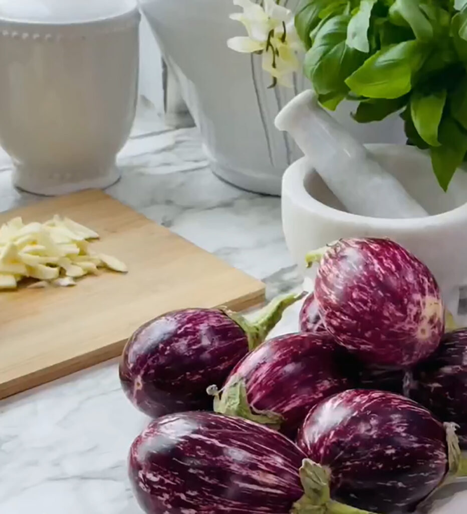 Cheesy Stuffed Eggplant in a Zesty Garlic Tomato Sauce, washed eggplants on countertop with a white mortar grinder next to a wooden chopping board with chopped garlic, fresh basil with two while canisters on a white countertop