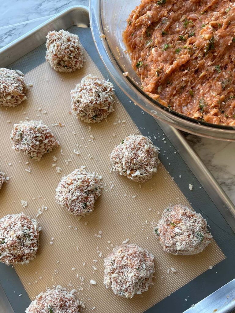 Cajun Chicken Meatball Pasta-crusted chicken meatballs on a baking sheet with a rubber baking mat with a glass bowl with seasoned ground chicken on a white countertop.