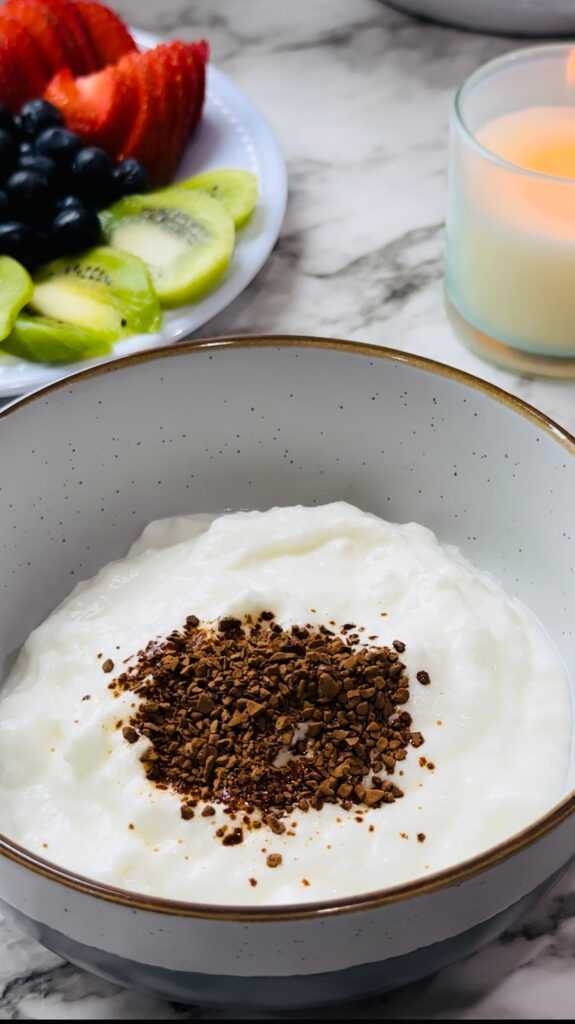 Healthy Mocha Coffee Yogurt yogurt and Instant Coffee together in a bowl with a plate of fruits and a lit candle in the background on a white countertop with white vases in the background.