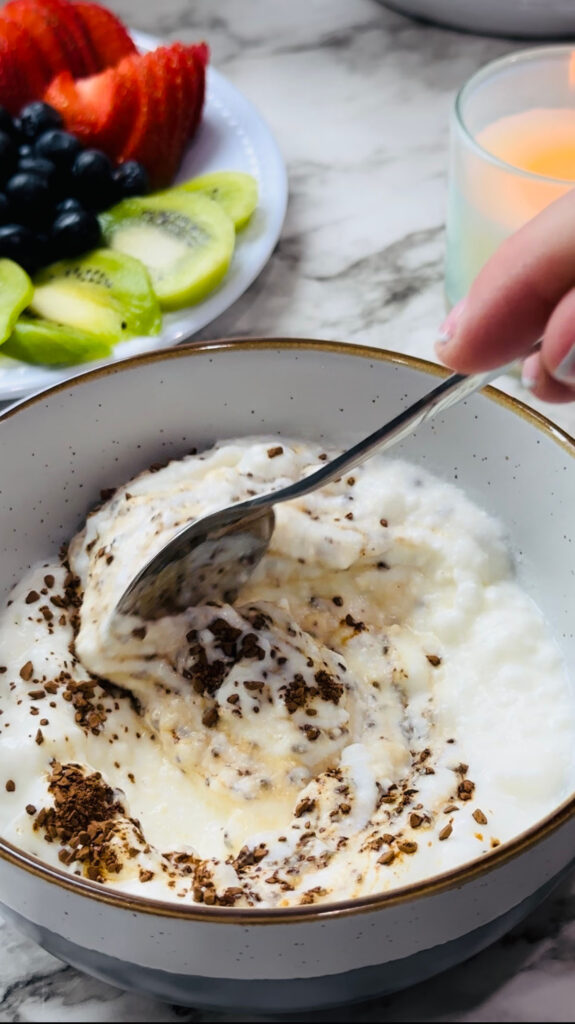 Healthy Mocha Coffee Yogurt yogurt and Instant Coffee mixed together in a bowl with a plate of fruits and a lit candle in the background on a white countertop with white vases in the background.