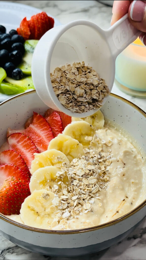 Healthy Mocha Coffee Yogurt yogurt with decorated strawberry and banana slices and cracked oats placed around the edge of the bowl with a plate of fruits and a lit candle in the background on a white countertop with white vases in the background.