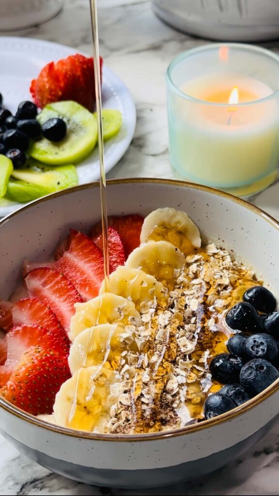 Healthy Mocha Coffee Yogurt yogurt with decorated strawberry and banana slices and cracked oats placed around the edge of the bowl and a drizzle of honey with a plate of fruits and a lit candle in the background on a white countertop with white vases in the background.