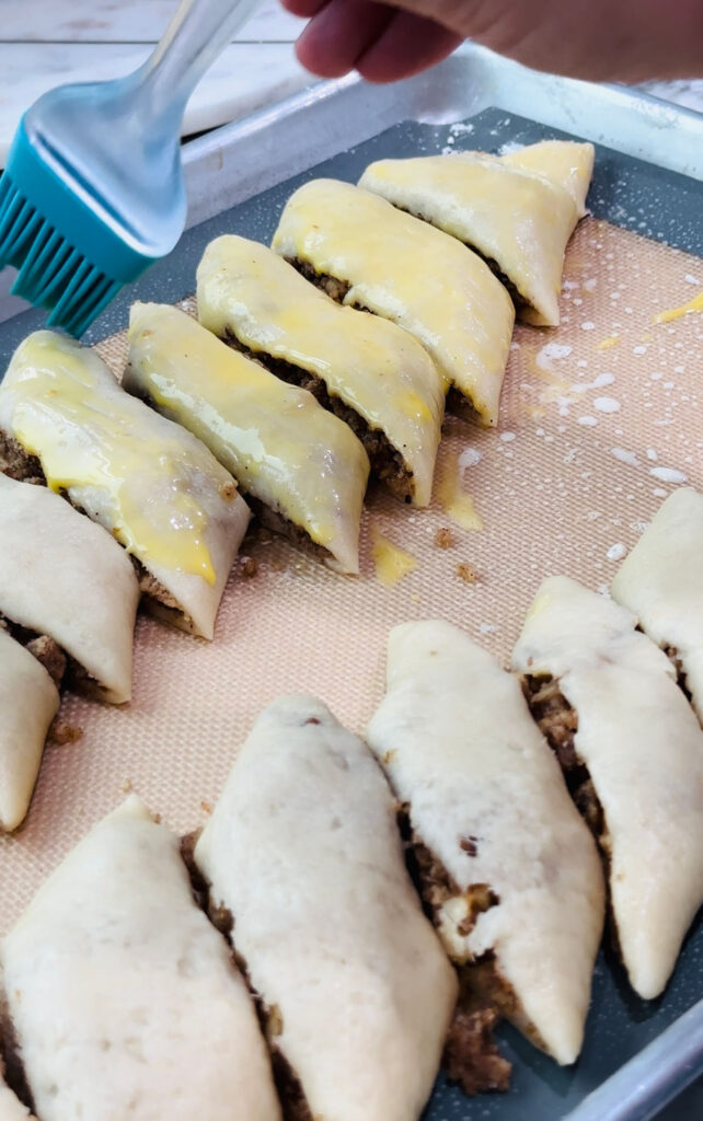 Walnut Roll Pastry cut pastry with egg wash brushed on top with a blue rubber cooking brush on a baking sheet lined with a rubber baking mat.
