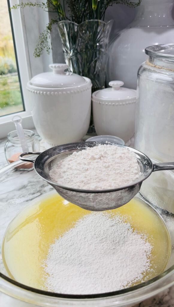Walnut Roll Pastry - placing sifted flour with a metal sifter of flour into a bowl of milk, oil, and eggs in a ceramic bowl in a white countertop in front of a kitchen window.