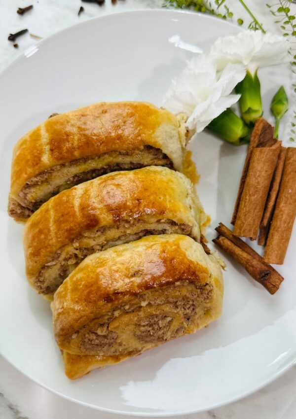 Walnut Roll Pastries on a white plate accented with cinnamon sticks and white carnations with cloves thrown on the table with greenery as accents around the plate on a white tabletop