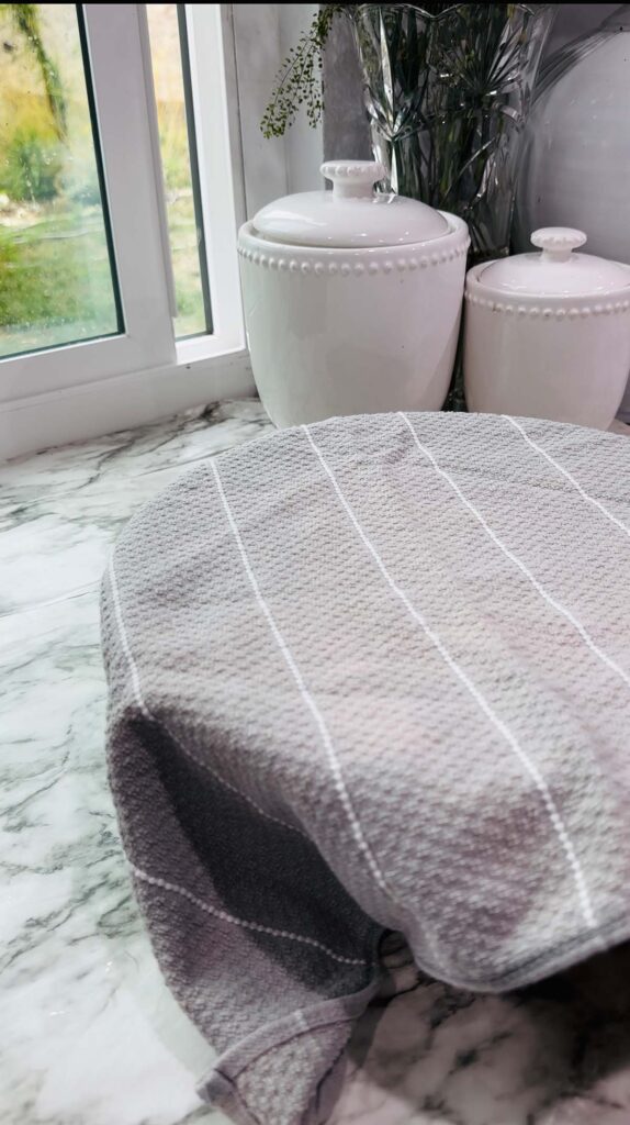 Walnut Roll Pastry - Glass bowl with dough with plastic wrap covered with a towel over dough with white canisters on a white granite countertop