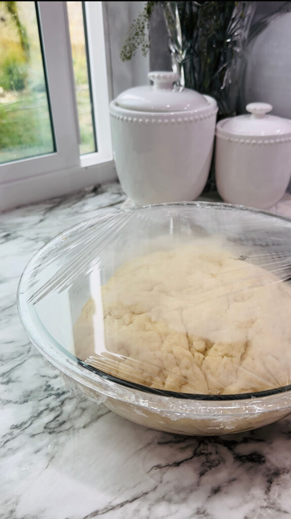 Walnut Roll Pastry - Glass bowl with dough with plastic wrap covered over dough with white canisters on a white granite countertop