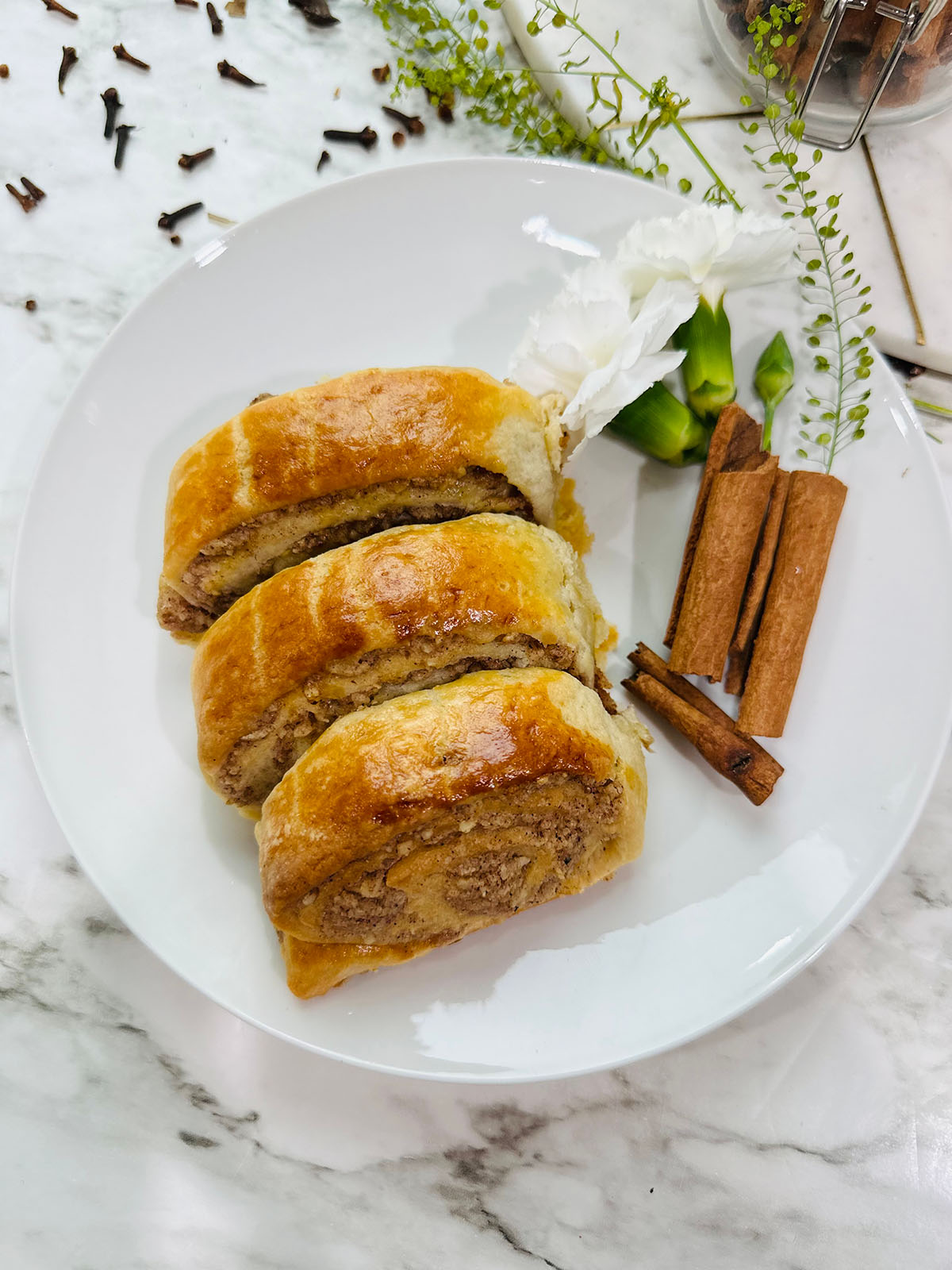 Three Walnut Rolls on a white plate with cinnamon sticks and white carnation with greenery accenting plate with cloves thrown on a white marble countertop